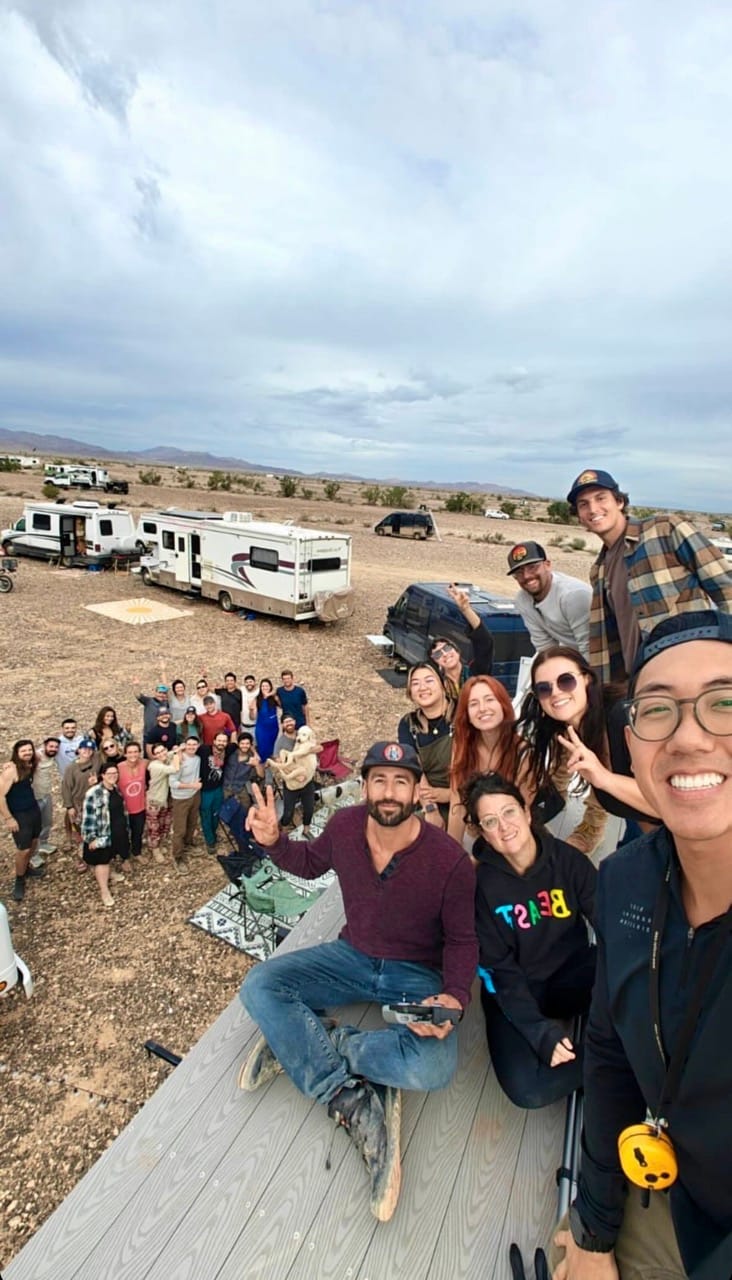 Two groups of vanlifers posing for a photo. One group sitting on a van roof with the second group in the background.