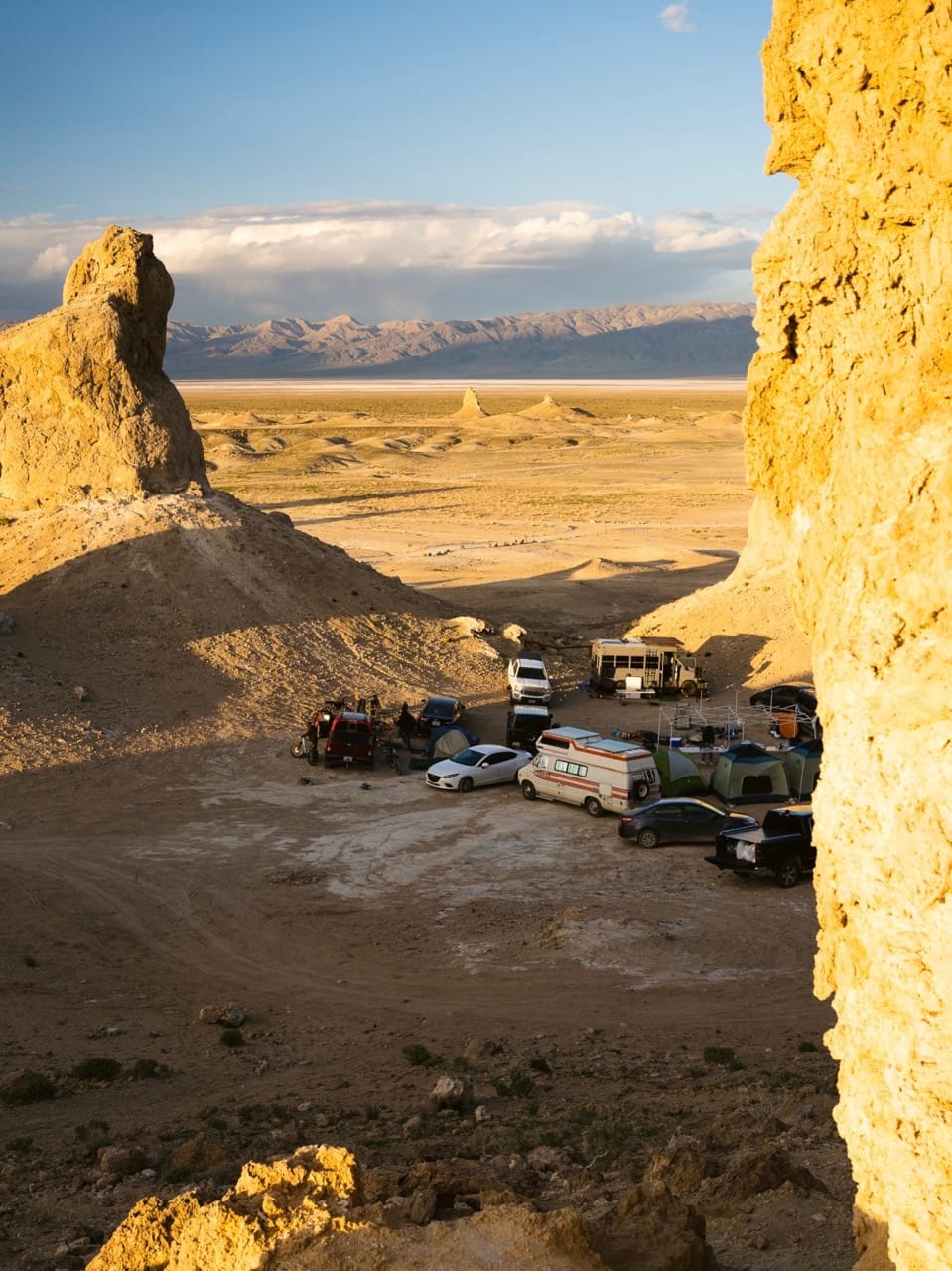 Several cars and campervans parked below a clip in a desert landscape with mountains in the distance