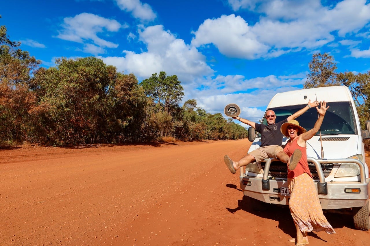 Vanlife couple posing enthusiastically in front of their van on a dusty road in the middle of nowhere